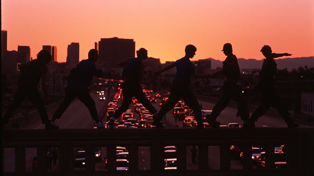 Dancers in silhouette on an overpass rail, city traffic and a sunset behind them
