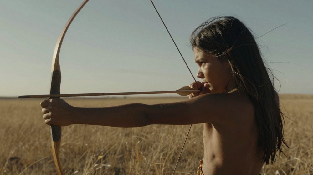 A Comanche boy at full draw on the open plain in daylight.